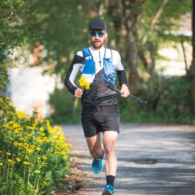 Maschsee Hannover - Ein Mann in Sportkleidung läuft auf einem gepflasterten Weg, der von Grün und gelben Blumen umgeben ist. Er nimmt am Hannover Triathlon teil, trägt eine blaue Trinkweste, eine Sonnenbrille und eine Kappe und hält einen Strauß gelber Blumen in der Hand, während er vorwärts rennt. - maschseefest Hannover