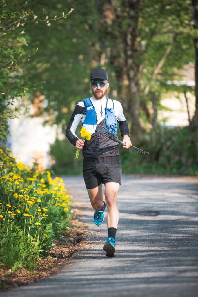 Maschsee Hannover - Ein Mann in Sportkleidung läuft auf einem gepflasterten Weg, der von Grün und gelben Blumen umgeben ist. Er nimmt am Hannover Triathlon teil, trägt eine blaue Trinkweste, eine Sonnenbrille und eine Kappe und hält einen Strauß gelber Blumen in der Hand, während er vorwärts rennt. - maschseefest Hannover