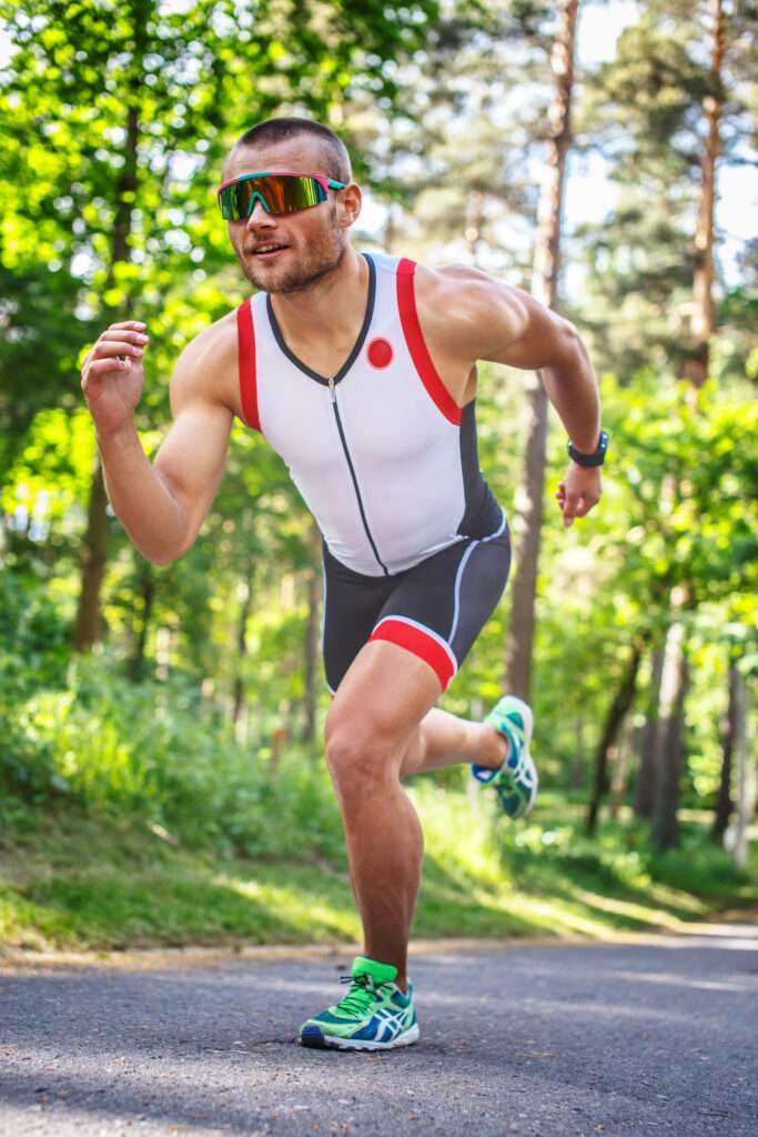 Maschsee Hannover - Ein Mann in Sportkleidung läuft auf einem gepflasterten Weg durch ein Waldstück, vielleicht trainiert er für den Hannover Triathlon. Er trägt ein weiß-schwarzes ärmelloses Oberteil mit roten Akzenten, schwarze Shorts, grüne Laufschuhe und eine verspiegelte Sonnenbrille. Üppige Bäume und Sonnenlicht rahmen die Szene ein. - maschseefest Hannover