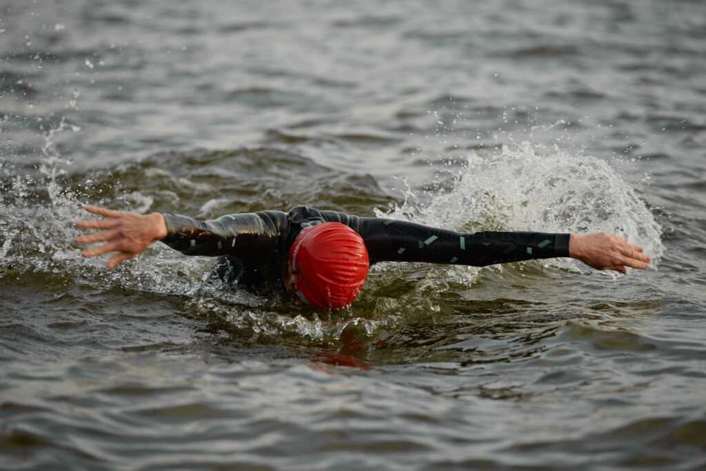Maschsee Hannover - Inmitten der dunklen, unruhigen Gewässer des Hannover Triathlons vollführt ein Schwimmer mit roter Schwimmkappe und schwarzem Neoprenanzug einen kraftvollen Freistilzug. Spritzer spritzen um ihn herum, während seine Arme durch das Wasser gleiten, und verkörpern die Entschlossenheit, die dieses berühmte Event weckt. - maschseefest Hannover