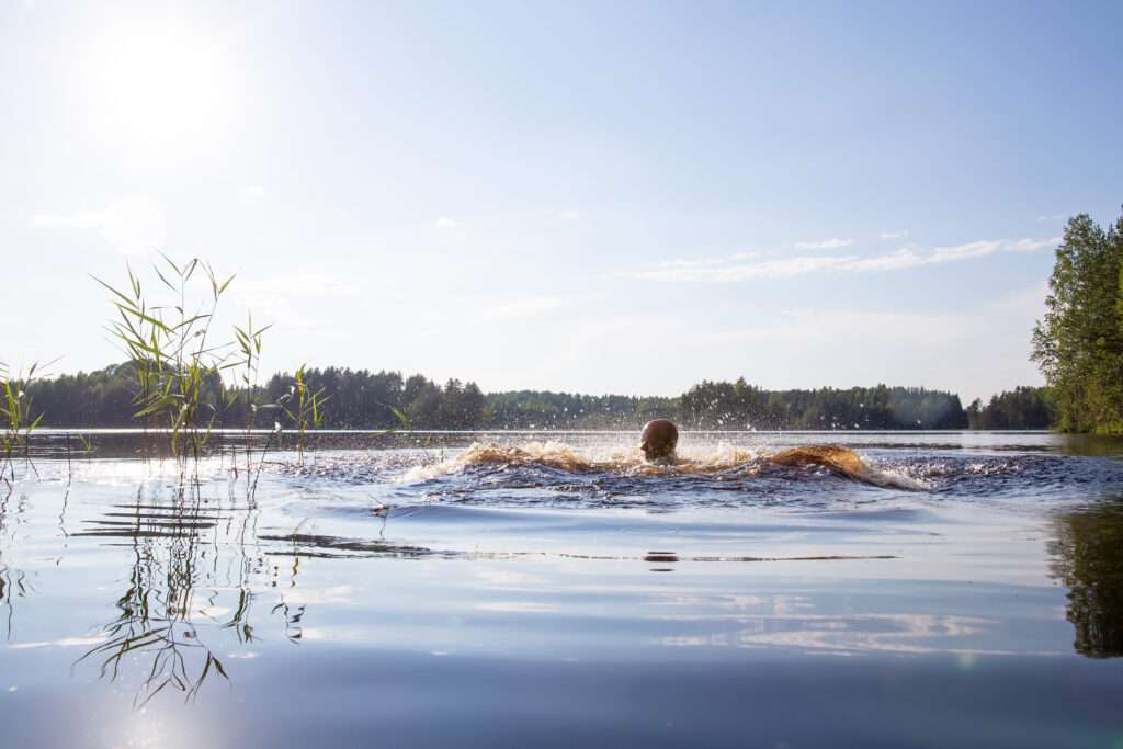 Maschsee Hannover - Eine Person schwimmt in einem ruhigen See. Um sie herum bilden sich Wellen wie beim Start eines Hannover-Triathlons. Die Sonne scheint hell am klaren blauen Himmel. Hohes Gras säumt den Wasserrand auf der linken Seite und dichte Bäume bilden eine ruhige Kulisse. - maschseefest Hannover
