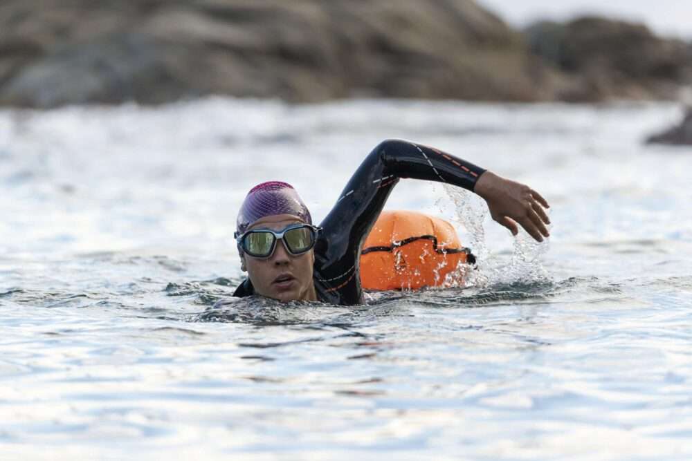 Maschsee Hannover - Eine Person, die am Hannover Triathlon teilnimmt, trägt Neoprenanzug, Badekappe und Schwimmbrille und schwimmt im Freistil im offenen Wasser. Ein orangefarbenes Schwimmgerät schleift hinter ihr her, während sie an felsigen Landformen vorbeigleitet, die im Hintergrund teilweise sichtbar sind. - maschseefest Hannover