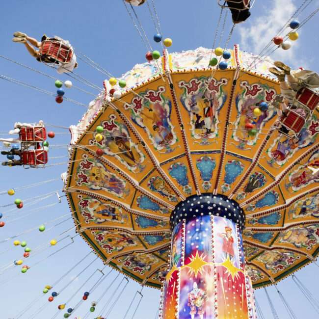 Maschsee Hannover - Auf einem farbenfrohen Karussell beim Schützenfest Hannover sitzen Menschen auf Stühlen, die an Metallketten befestigt sind. Das Karussell bietet lebendige Muster und Bilder vor einem blauen Himmel mit vereinzelten Wolken, während kugelähnliche Ornamente anmutig von oben herab schwingen. - maschseefest Hannover