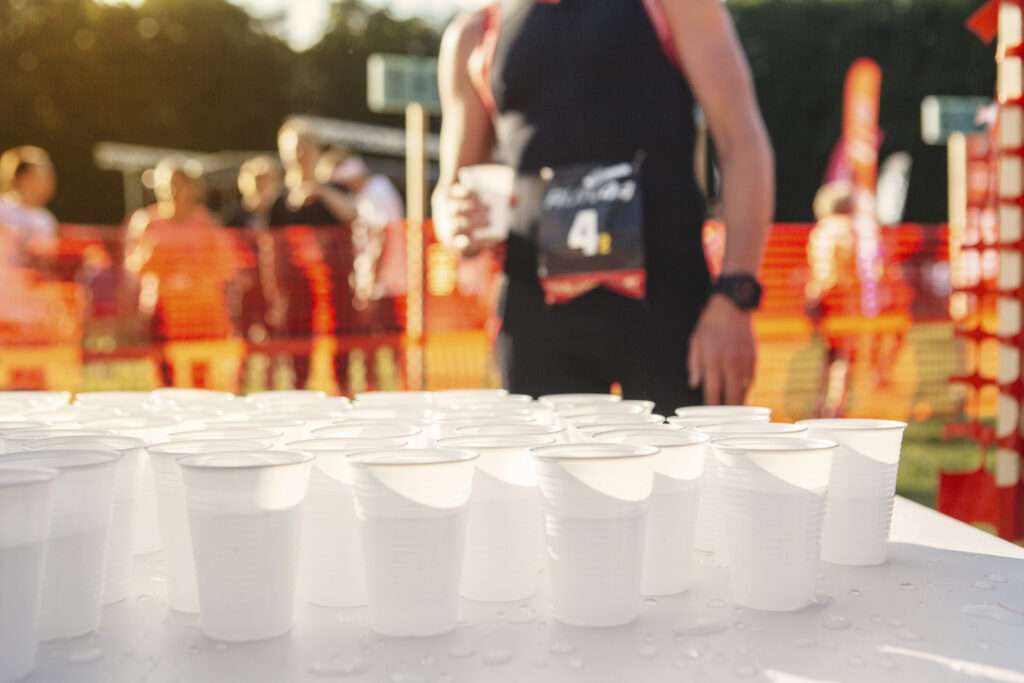 Maschsee Hannover - Reihen von mit Wasser gefüllten Plastikbechern säumen den Tisch beim Hannover Triathlon. Im Hintergrund hält ein Athlet mit nummerierter Startnummer einen Becher neben einem orangefarbenen Sicherheitszaun und genießt den sonnigen Tag im Freien bei diesem aufregenden Sportereignis. - maschseefest Hannover