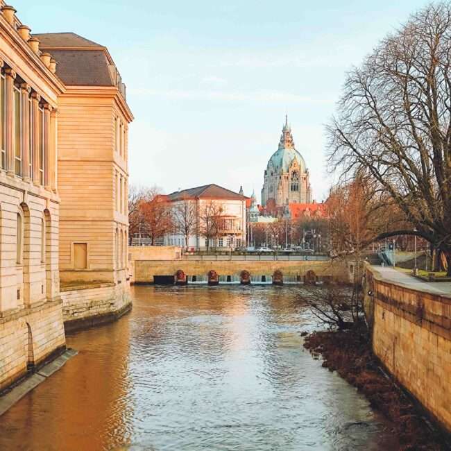Maschsee Hannover - Fluss mit Gebäuden auf beiden Seiten, im Hintergrund eine große Kuppel. Bäume mit spärlichem Laub säumen das Flussufer. Die Szene spielt unter klarem Himmel, warmes Sonnenlicht wirft weiche Schatten. - maschseefest Hannover
