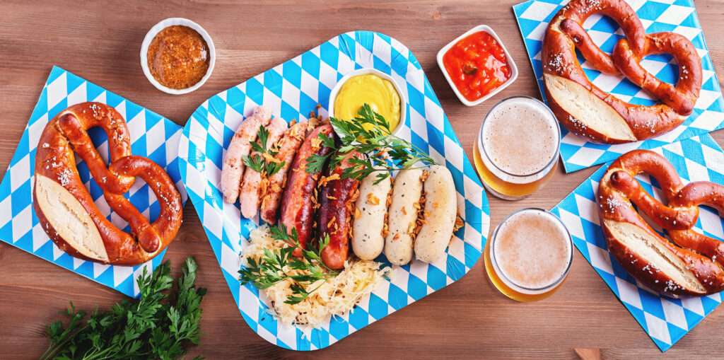 Maschsee Hannover - Ein Buffet mit Brezeln, Würstchen, Sauerkraut und Gewürzen auf blau-weiß karierten Tellern, das an die Hannoveraner Wiesn erinnert. Dazu gibt es Biergläser und Petersilie, alles elegant angerichtet auf einem rustikalen Holztisch. - maschseefest Hannover