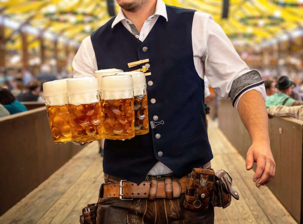 Maschsee Hannover - Eine Person in traditioneller Kleidung trägt auf der lebhaften Hannoveraner Wiesn in jeder Hand mehrere große Bierkrüge. Die Kulisse ist festlich: Ein gelb-weiß gestreiftes Zeltdach und Menschen im Hintergrund sitzen an Tischen und genießen die ausgelassene Stimmung. - maschseefest Hannover