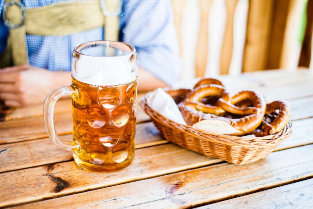 Maschsee Hannover - Ein großer Glaskrug Bier steht auf einem Holztisch neben einem Weidenkorb mit Brezeln und einem Stück Papier und fängt die festliche Atmosphäre der Hannoveraner Wiesn ein. Im Hintergrund ist eine Person in einem blaukarierten Hemd teilweise zu sehen, was zur ausgelassenen Atmosphäre beiträgt. - maschseefest Hannover