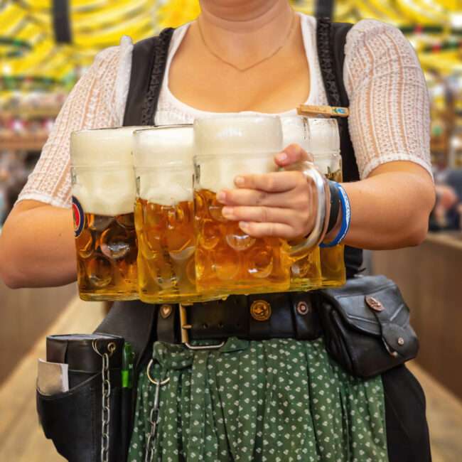 Maschsee Hannover - Eine Person in traditioneller Kleidung trägt auf der pulsierenden Hannoveraner Wiesn in jeder Hand mehrere große Bierkrüge mit Schaumkronen. Im Hintergrund herrscht ein geschäftiges Treiben auf dem Fest, über dem bunte Dekorationen wehen. - maschseefest Hannover