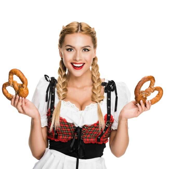Maschsee Hannover - Eine Person mit geflochtenen Haaren hält zwei Brezeln in der Hand und trägt ein traditionelles bayerisches Outfit mit rotem Karomuster und schwarzem Mieder über einer weißen Bluse, bereit für die Hannover Wiesn. Der Hintergrund ist schlicht weiß. - maschseefest Hannover