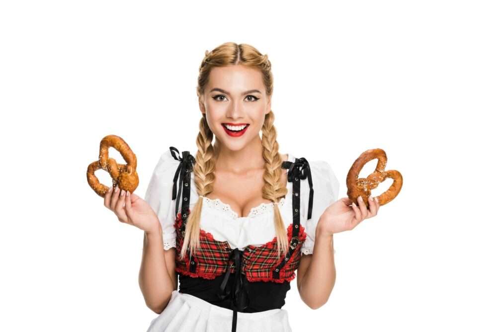 Maschsee Hannover - Eine Person mit geflochtenen Haaren hält zwei Brezeln in der Hand und trägt ein traditionelles bayerisches Outfit mit rotem Karomuster und schwarzem Mieder über einer weißen Bluse, bereit für die Hannover Wiesn. Der Hintergrund ist schlicht weiß. - maschseefest Hannover