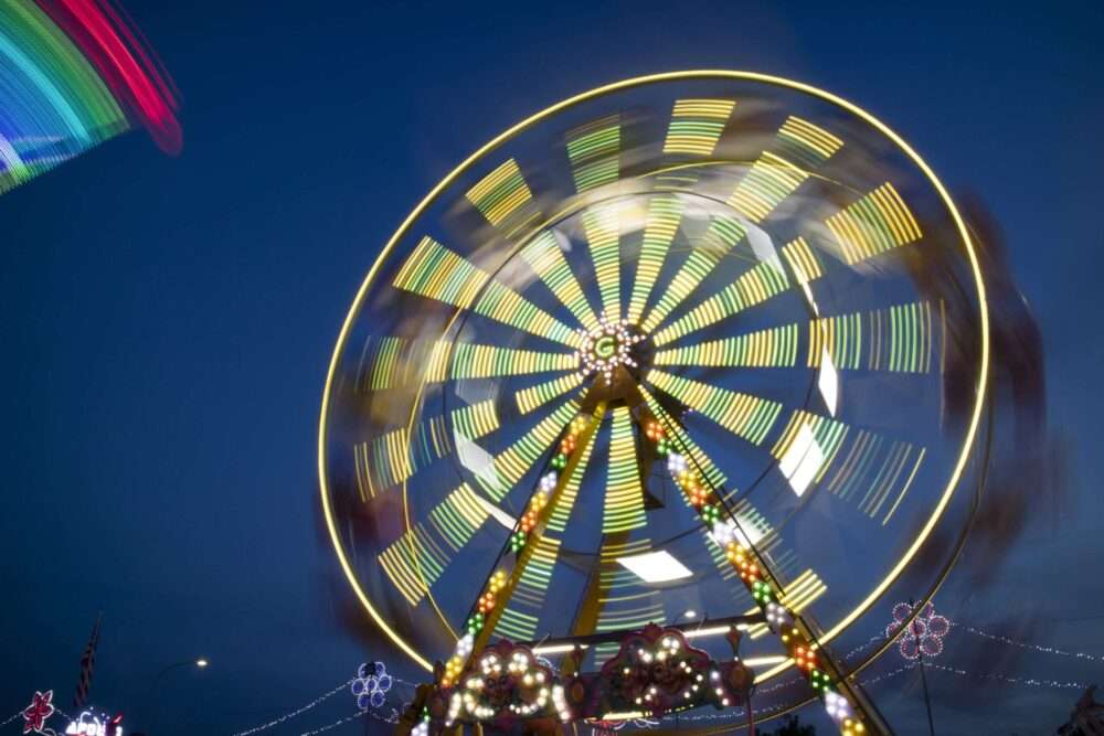 Maschsee Hannover - Ein hell erleuchtetes Riesenrad dreht sich in der Dämmerung auf dem Oktoberfest Hannover. Bunte Lichter erzeugen eine kreisförmige Bewegungsunschärfe. Der sich verdunkelnde Himmel bildet die Kulisse, während zusätzliche Lichter und Formen im Hintergrund funkeln und die lebendige Jahrmarktsatmosphäre einfangen. - maschseefest Hannover