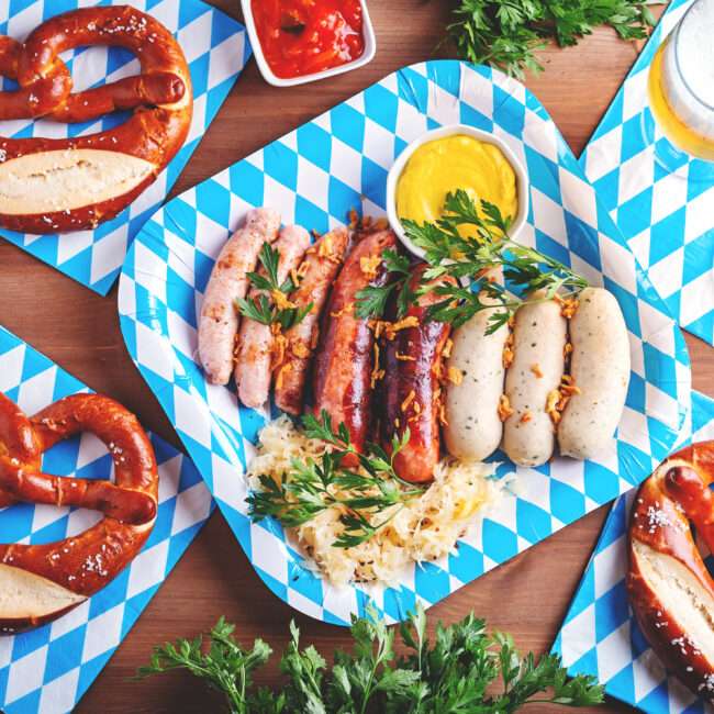 Maschsee Hannover - Auf einem Tisch mit blau-weiß karierten Tellern und Servietten im bayerischen Stil stehen Brezeln, verschiedene Würstchen mit Petersiliengarnitur, Sauerkraut, Senf, eine Schüssel körnigen Senf und zwei Gläser Bier – eine perfekte Kulisse für eine Oktoberfestfeier, die an die Festlichkeiten in Hannover erinnert. - maschseefest Hannover