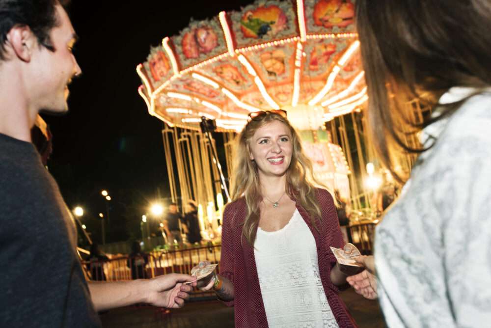 Maschsee Hannover - Eine Gruppe von Menschen steht neben einem hell erleuchteten Karussell auf dem Oktoberfest in Hannover. In der Mitte steht eine Frau in weißem Kleid und bordeauxrotem Pullover. Sie hält Geldscheine in der Hand und lächelt, während zwei andere ihr Geld reichen. Die Stimmung ist ausgelassen und festlich. - maschseefest Hannover