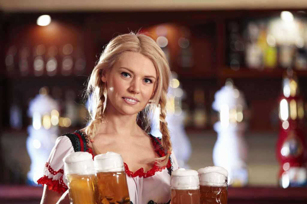 Maschsee Hannover - Eine Frau mit geflochtenen Haaren und traditioneller bayerischer Kleidung hält auf dem Oktoberfest in Hannover mehrere Krüge Bier in der Hand. Sie steht vor einer Bar, im Hintergrund sind unscharfe Regale mit Flaschen zu sehen. - maschseefest Hannover