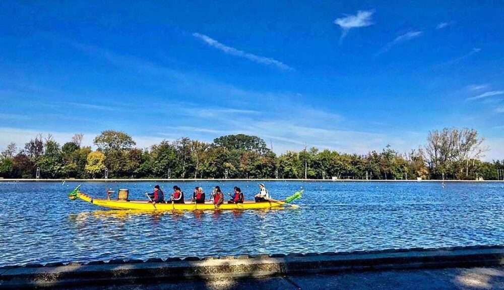 Maschsee Hannover - Ein langes gelbes Drachenboot gleitet über einen ruhigen, blauen See. Sechs Ruderer rudern beim Drachenbootrennen in Hannover in perfekter Harmonie. Die Ruderer, in rote Schwimmwesten gekleidet, werden von den Bäumen am Ufer unter einem klaren, blauen Himmel eingerahmt. - maschseefest Hannover
