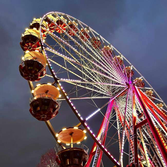 Maschsee Hannover - Beim Frühlingsfest 2025 in Hannover erstrahlt ein großes Riesenrad vor einem dunklen, bewölkten Himmel in bunten Lichtern. Die Konstruktion besteht aus mehreren Gondeln und filigranen Metallrahmen. Die Perspektive von unten unterstreicht die Größe und Erhabenheit des Rades. - maschseefest Hannover