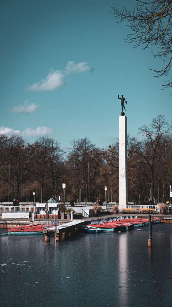 Maschsee Hannover - Eine hohe Marmorstatue steht auf einem Sockel in der Nähe einer Uferpromenade, an der kleine Boote angedockt sind. Bäume mit kahlen Ästen säumen den Hintergrund unter einem teilweise bewölkten Himmel. - maschseefest Hannover