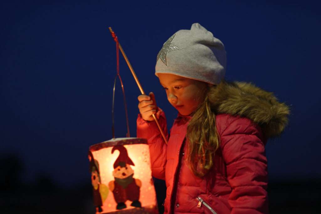 Maschsee Hannover - Ein junges Mädchen in rotem Mantel und grauem Hut hält eine leuchtende Laterne mit Zwergenmotiven in der Hand. Sie steht nachts im Freien und träumt vielleicht vom Oktoberfest in Hannover, während sie das Licht in ihrer rechten Hand aufmerksam betrachtet. - maschseefest Hannover