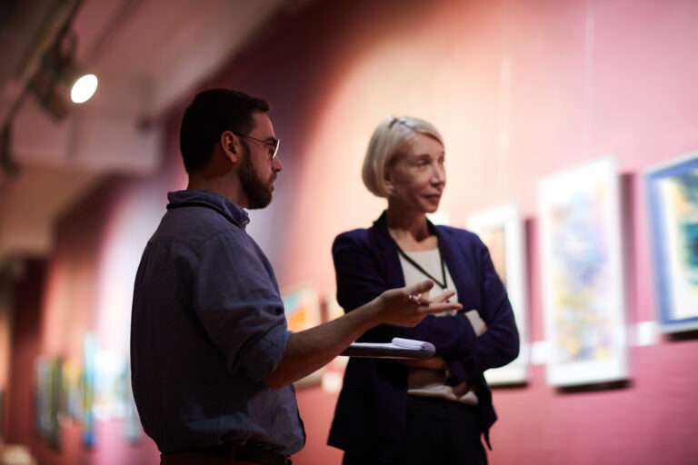 Maschsee Hannover - Ein Mann und eine Frau stehen in der schwach beleuchteten Kunstgalerie des Museums Hannover und unterhalten sich. Der Mann umklammert ein Notizbuch, während an der roten Wand hinter ihnen verschwommene Gemälde hängen, die durch ihre Beleuchtung ihren künstlerischen Reiz unterstreichen. - maschseefest Hannover