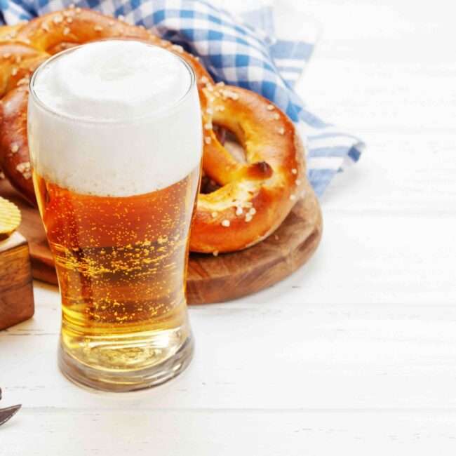 Maschsee Hannover - Ein Glas schaumiges Bier steht auf einer Holzfläche und erinnert an das Oktoberfest. Dazu gibt es ein Tablett mit geriffelten Kartoffelchips und Salzbrezeln. Im Hintergrund ist teilweise ein blau-weiß kariertes Tuch zu sehen, das den lebendigen Geist Hannovers heraufbeschwört. - maschseefest Hannover