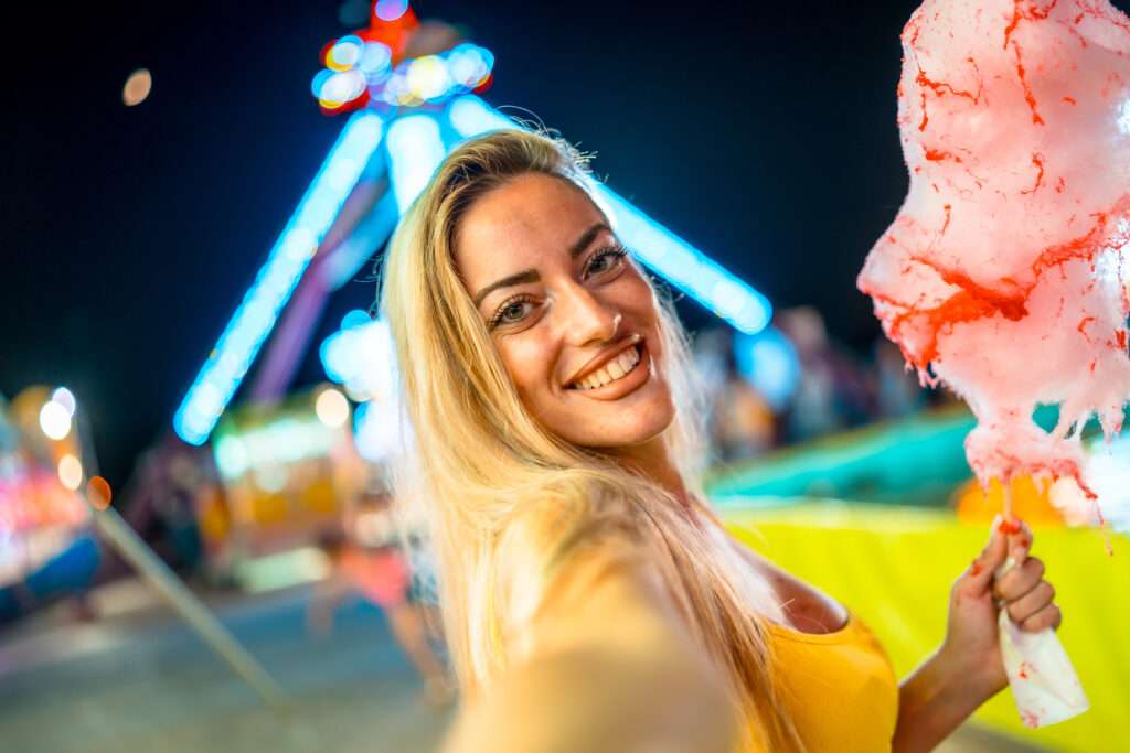 Maschsee Hannover - Eine lächelnde Person mit langen blonden Haaren macht ein Selfie mit rosa Zuckerwatte in der Hand beim Schützenfest Hannover. Sie befinden sich nachts in einem Vergnügungspark, im Hintergrund glitzern hell erleuchtete Fahrgeschäfte. - maschseefest Hannover