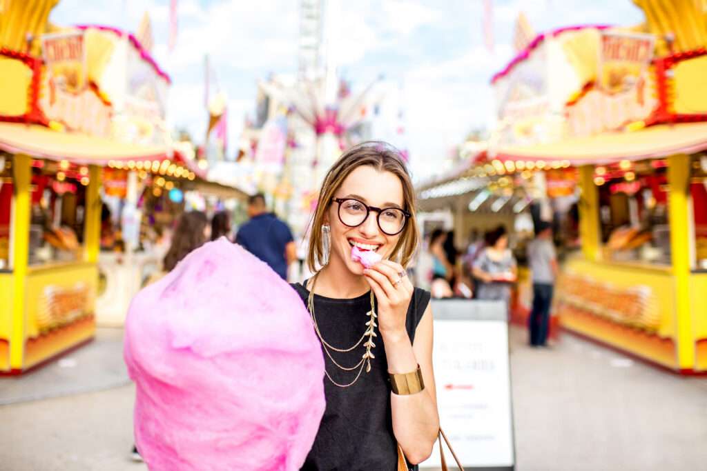 Maschsee Hannover - Eine Person mit Brille und schwarzem Oberteil lächelt und hält rosa Zuckerwatte in der Hand, während sie vor den leuchtenden Lichtern und geschäftigen Essensständen des Schützenfests Hannover steht. - maschseefest Hannover