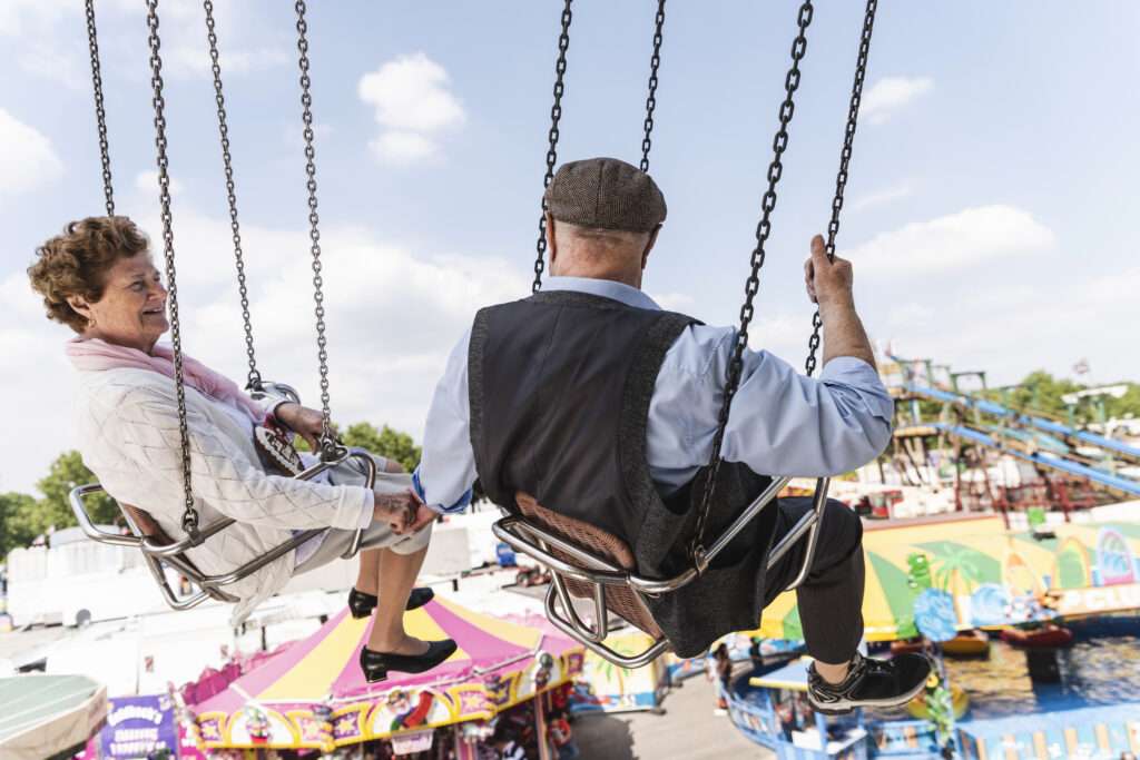 Maschsee Hannover - Zwei ältere Personen sitzen auf einem Kettenkarussell beim Schützenfest Hannover. Der Mann trägt Mütze und Weste, die Frau einen leichten Mantel und einen Schal. Unter ihnen sind bunte Fahrgeschäfte und Zelte zu sehen. Der Himmel ist teilweise bewölkt. - maschseefest Hannover