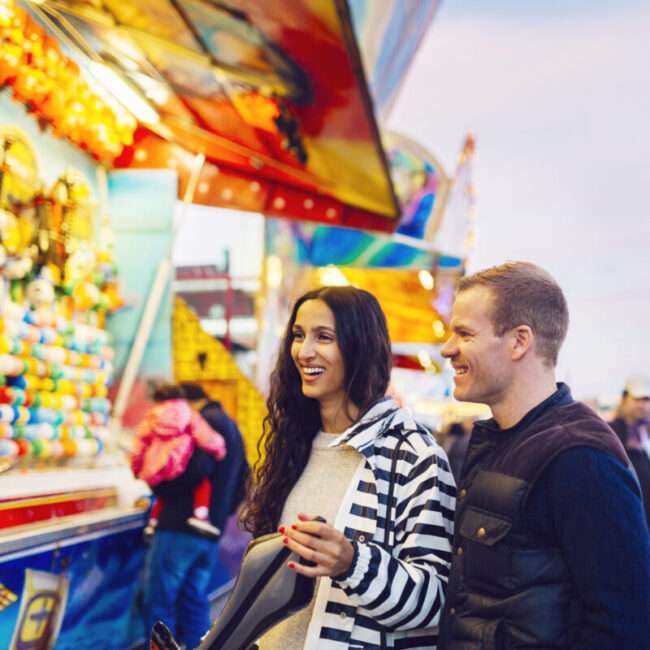 Maschsee Hannover - Ein Mann und eine Frau stehen lächelnd vor einem bunten Karnevalsspielstand voller Plüschtiere auf dem Schützenfest Hannover. Der Himmel ist bewölkt und strahlt in einem warmen Licht, während Menschen, darunter auch Kinder, in der festlichen Kulisse umherschlendern. - maschseefest Hannover